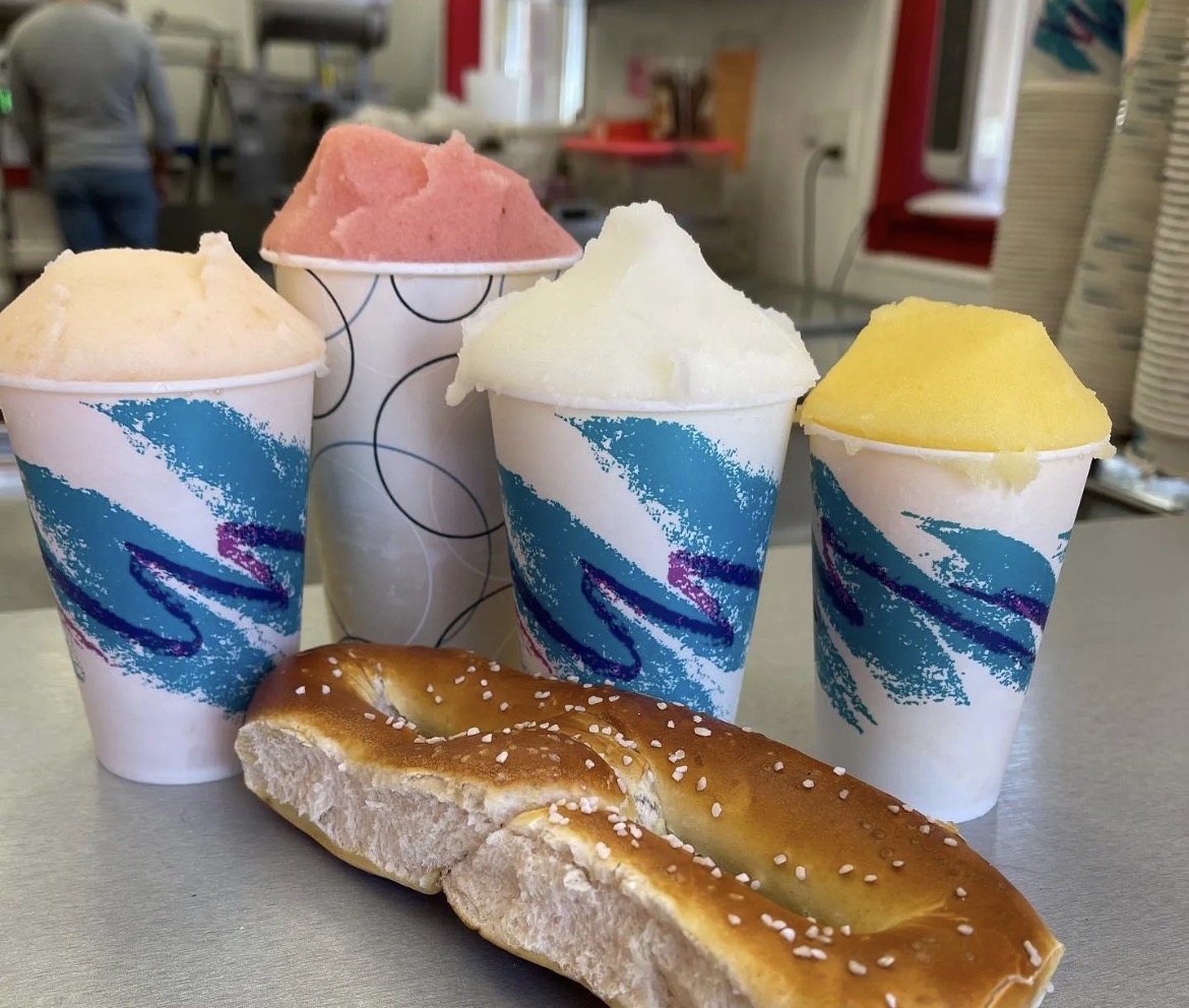 Four cups of water ice in cherry, lemon, mango and vanilla flavors behind a salted soft pretzel on a steel counter