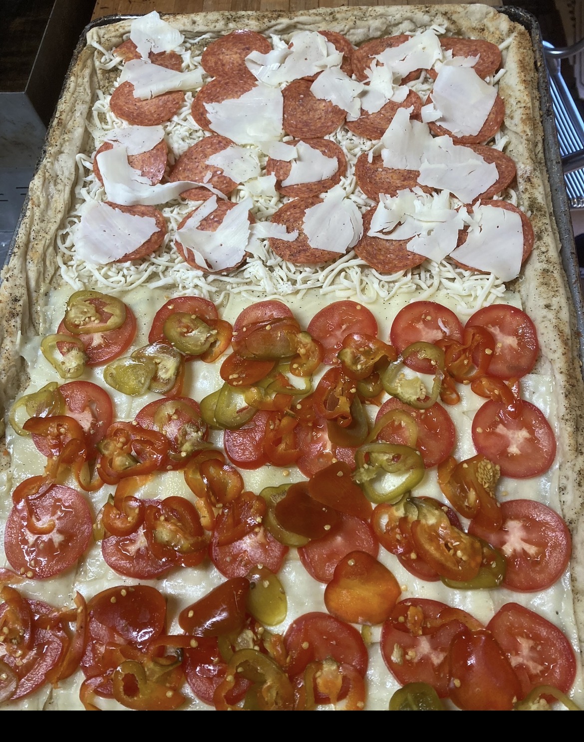 Rectangular sheet-pan pizza half topped with pepperoni and shaved cheese, half with sliced tomatoes and hot and sweet peppers